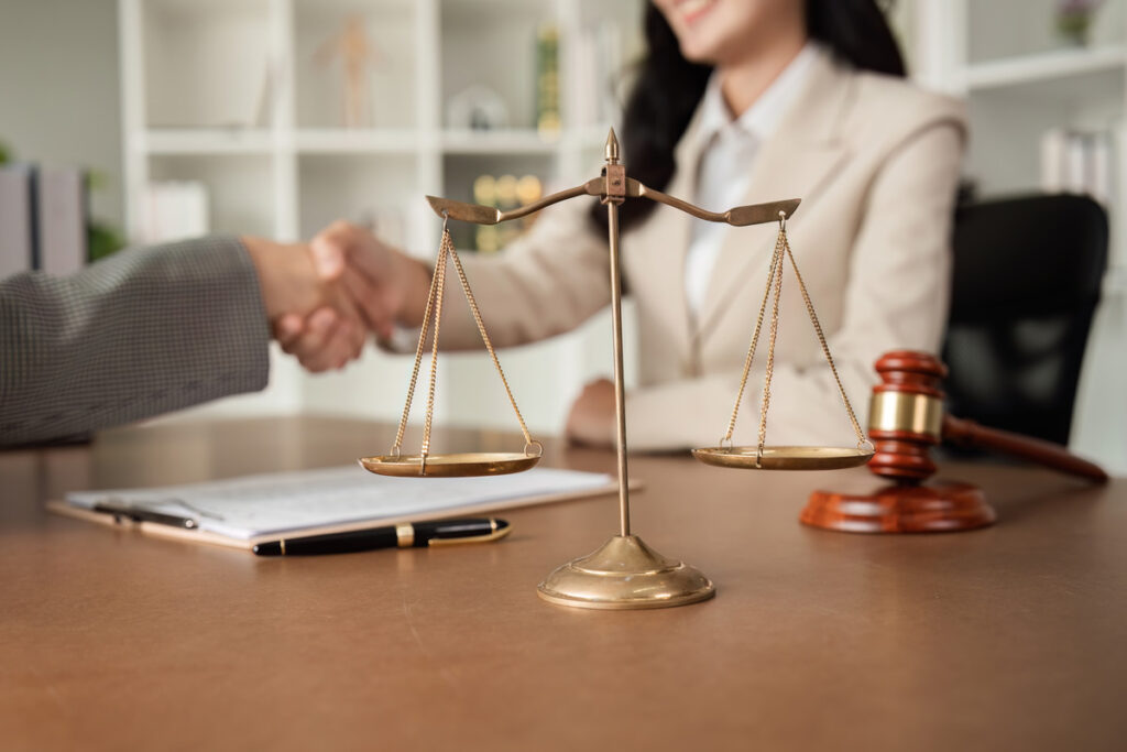 Close-up of a balanced scale, a gavel, and a pen on a desk, illustrating how long does a divorce take. Two people shaking hands in the background signify an agreement in a legal or professional setting