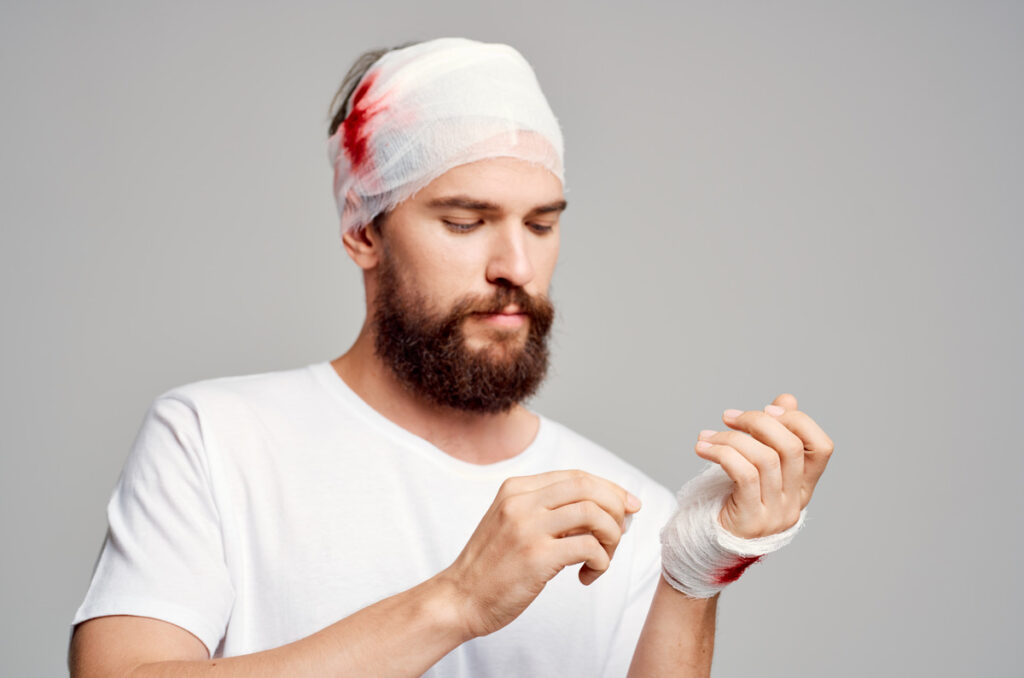 Person with blood-stained bandages on head and wrist adjusting bandage on gray background, illustrating the physical impact of assault and battery