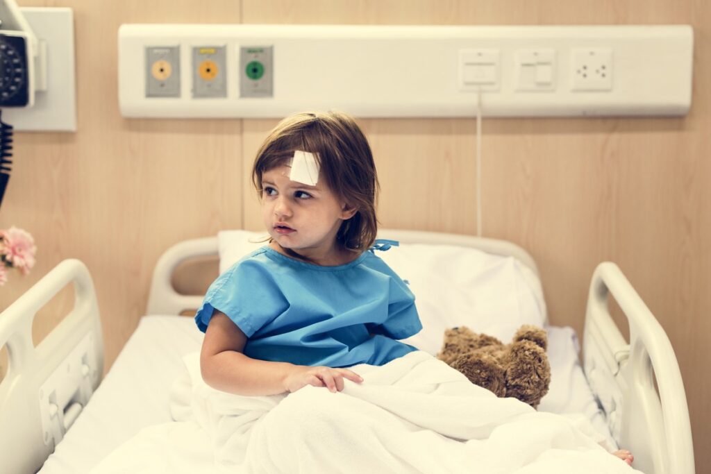 Child sitting on a hospital bed, wearing a blue hospital gown, with a teddy bear beside them, illustrating the role of guardianship in making critical healthcare decisions and how it might override parental rights