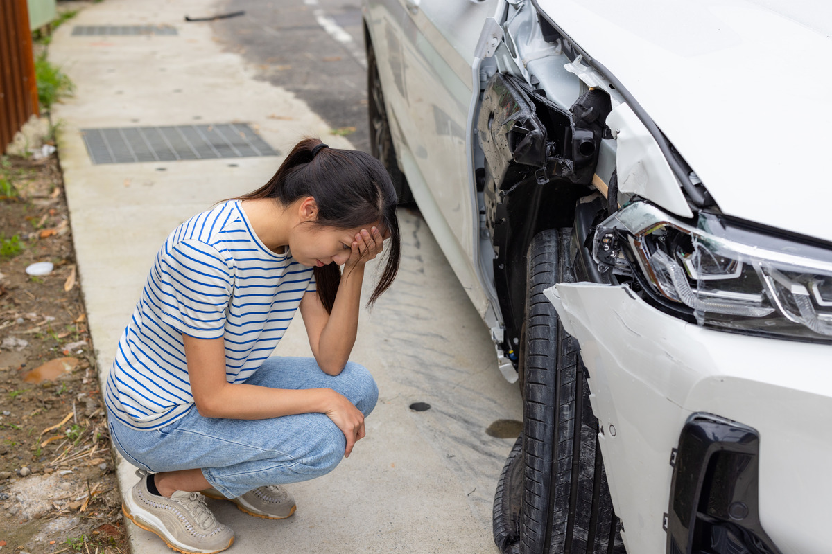 A woman kneels by a white car with visible damage, pondering the reasons behind the slow car accident settlement process