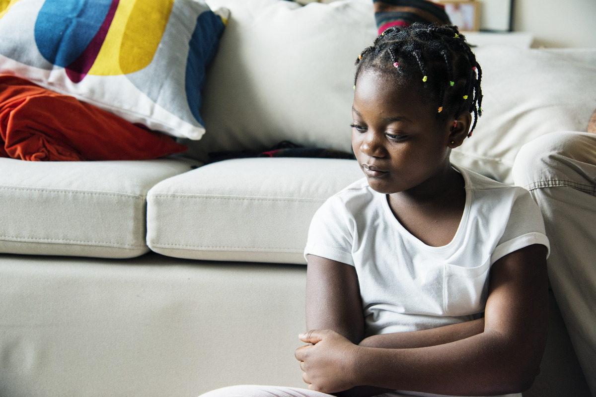 A young girl with braided hair sits on a beige couch, looking thoughtful. A colorful pillow is visible in the background