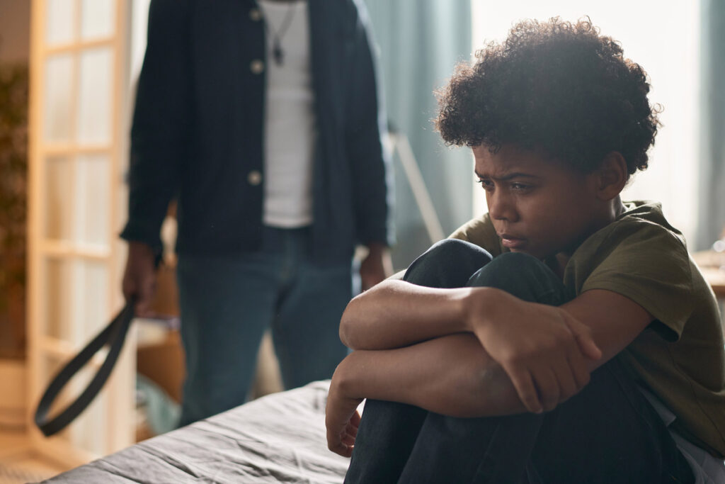 A child sits on a bed with knees hugged to their chest, appearing upset, while an adult stands in the background holding a belt