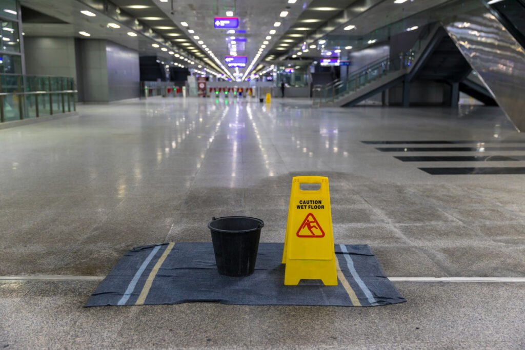 A wet floor sign in an airport warns of slippery conditions, reminding to report slips and seek assistance after an accident
