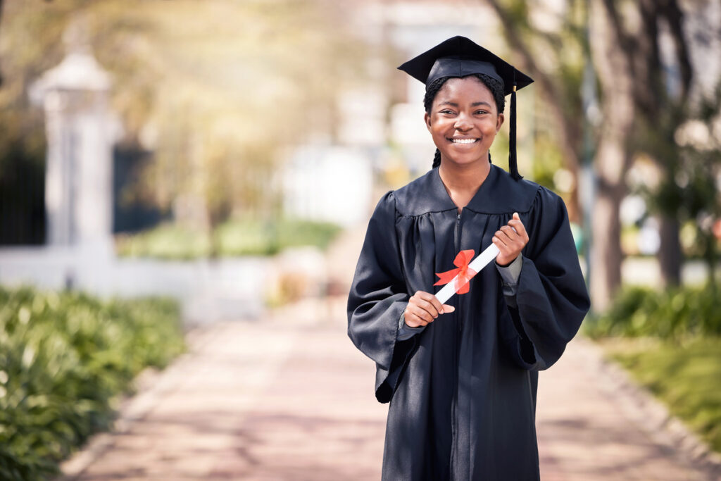 A young woman in a graduation gown proudly holds a red ribbon, symbolizing her academic achievement