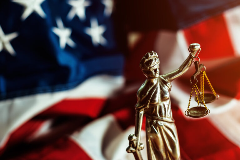 Scales of justice statue in front of a blurred American flag, symbolizing law and justice in the United States